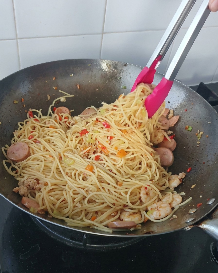 中華鍋から、海老やスライスソーセージが混ざったたっぷりのスパゲティを、ピンクの持ち手のトングで持ち上げている様子。