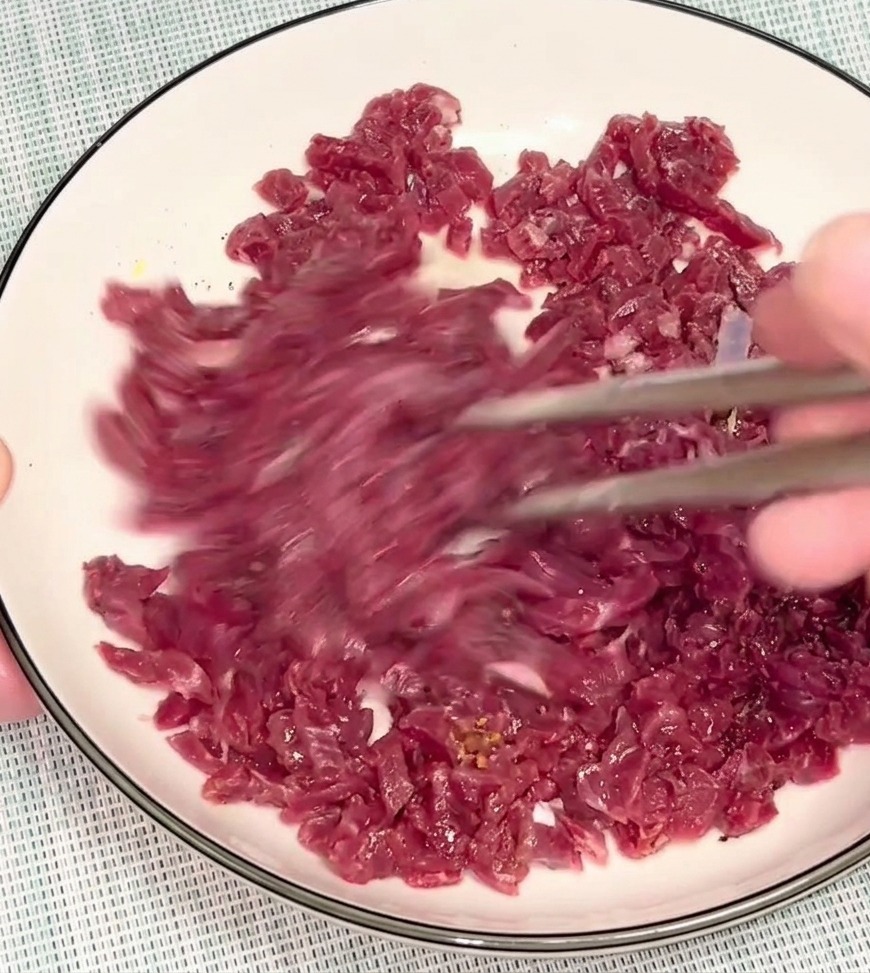 銀の箸を使って、白いボウルの中で生の赤身のひき肉と調味料をしっかりと混ぜ合わせている様子。