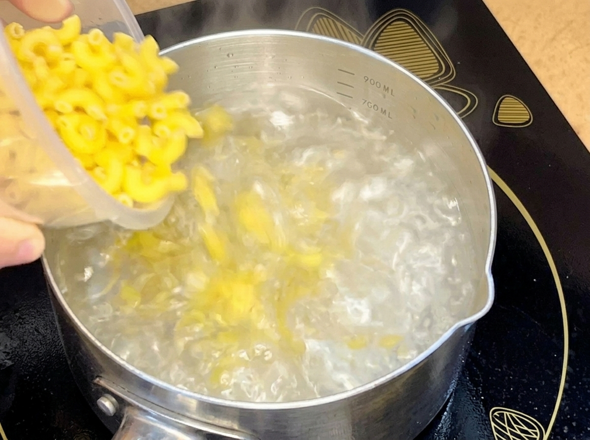 ステンレスの鍋で沸騰したお湯の中に、容器から乾燥したエルボーマカロニを投入している様子。