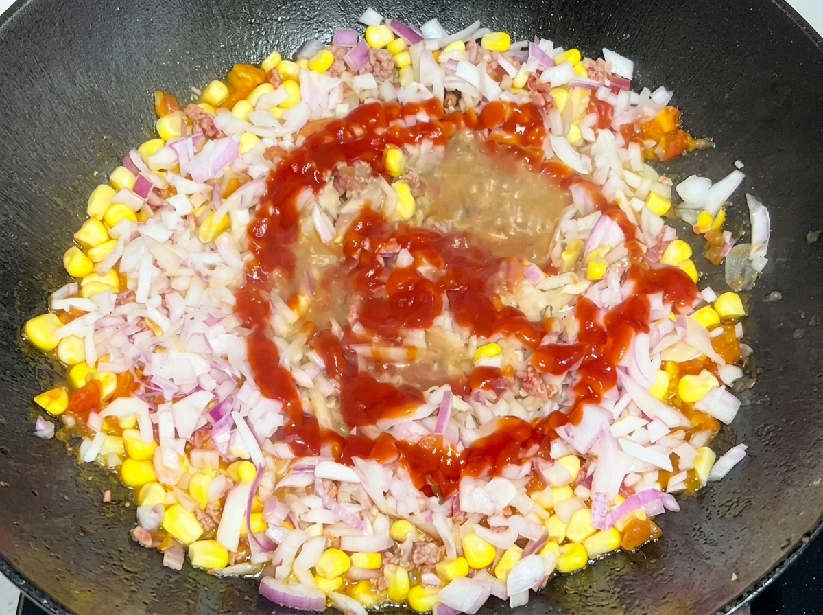 フライパンの中の牛肉とコーンの具材に、細かく刻んだ赤玉ねぎとケチャップを回し入れている様子。