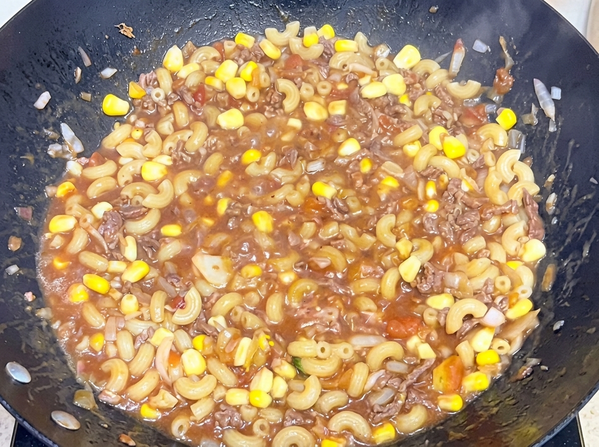 大きな中華鍋の中で、茹で上がったマカロニを旨味たっぷりの牛肉トマトソースにしっかりと和えている様子。