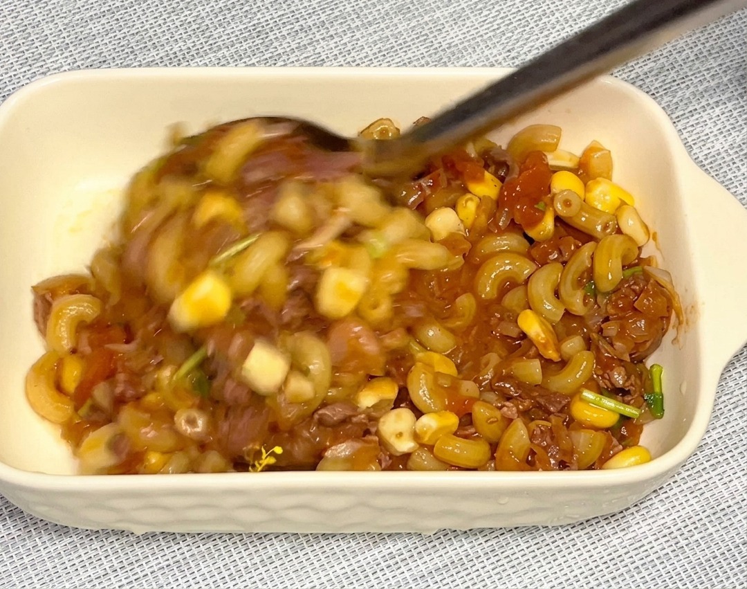 大きなメタルスプーンを使い、コーンや牛肉が入った具だくさんのマカロニを長方形の白いグラタン皿に移し替えている様子。