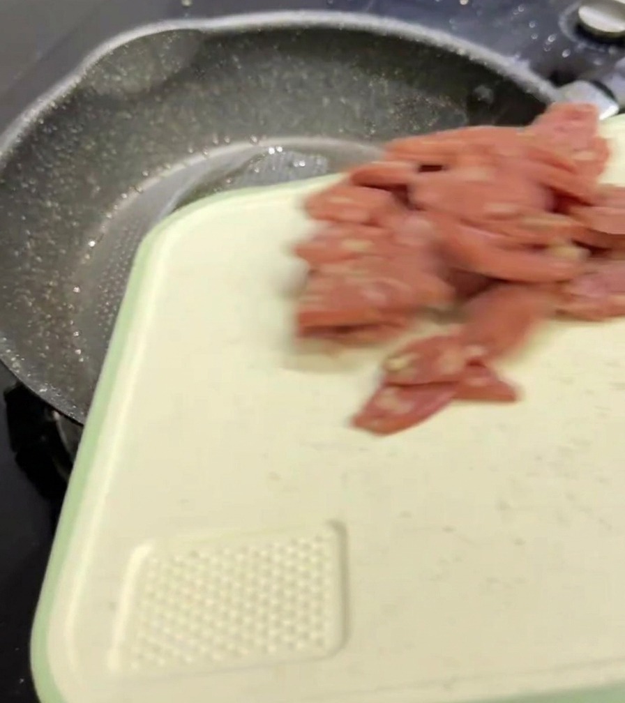 白いまな板からフライパンの中へ、スライスされたピンク色のサラミを投入している様子。