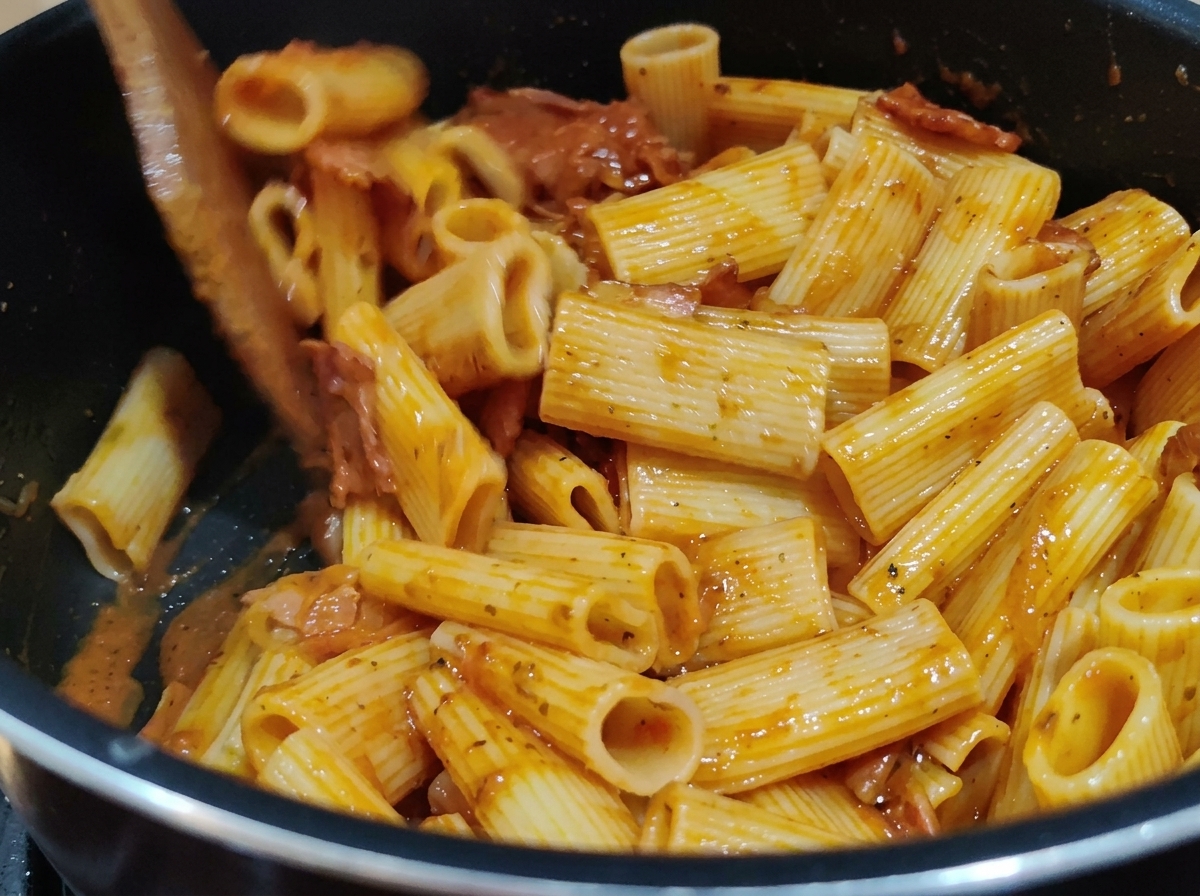 鍋の中で木べらを使い、リガトーニパスタに濃厚な赤色のトマトソースをしっかりと絡めている様子