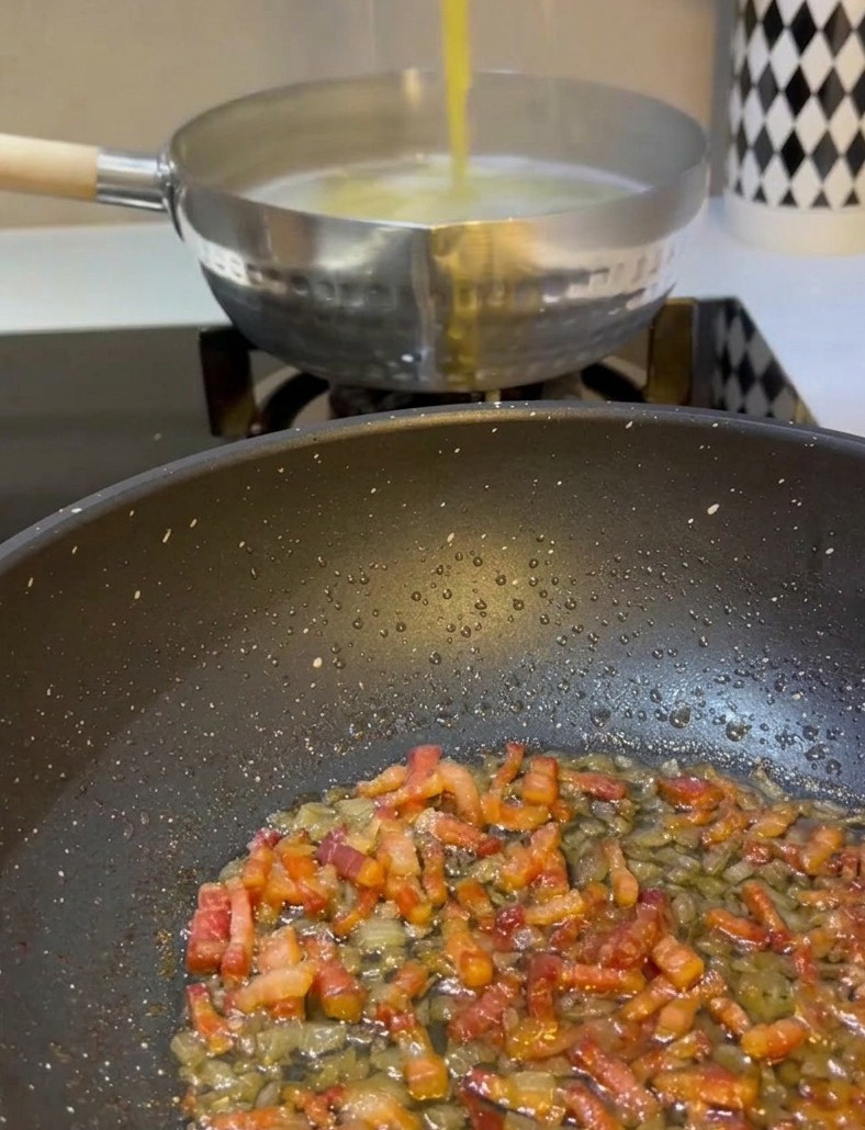 トングを使い、沸騰した鍋から茹で上がった黄色のスパゲッティを持ち上げ、炒めたパンチェッタと玉ねぎが入ったフライパンへ直接移している様子。