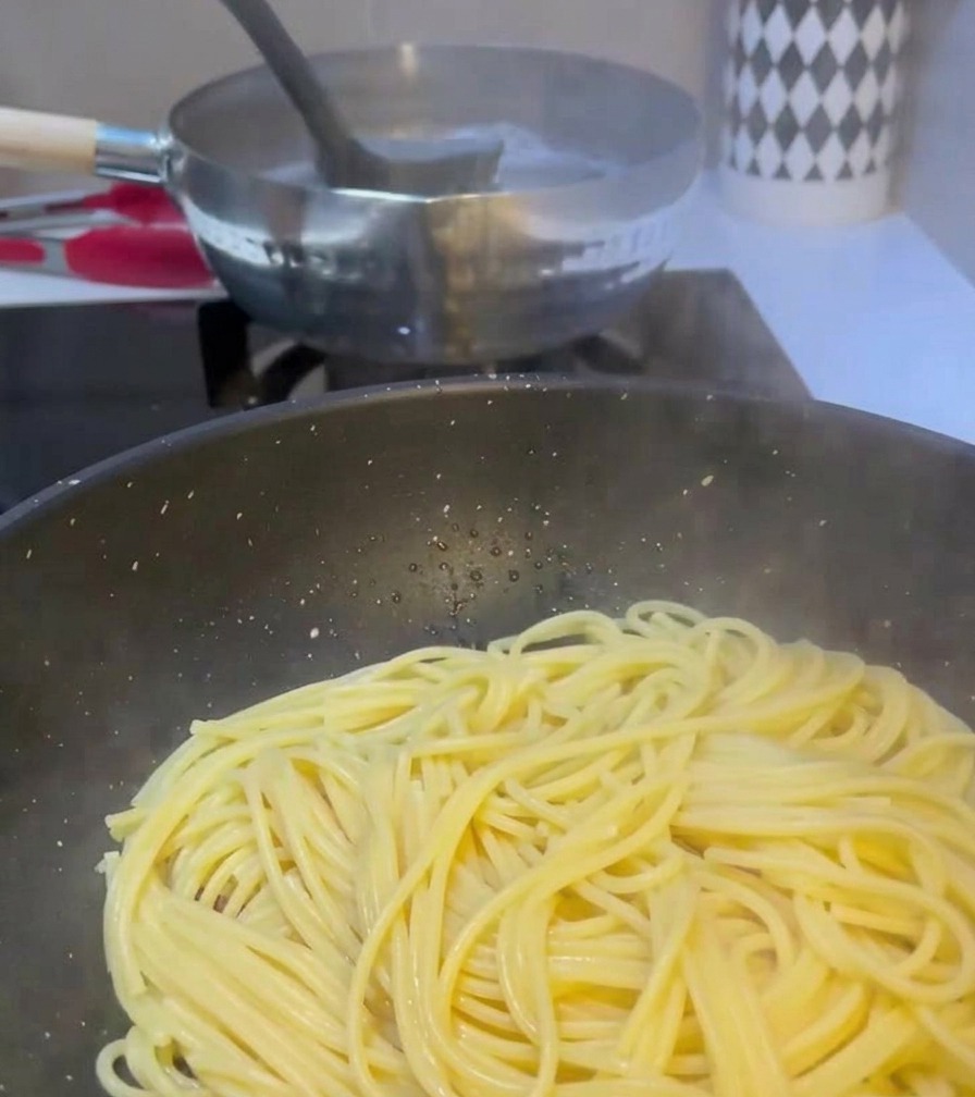 沸騰したパスタ鍋からお玉で白く濁った茹で汁をすくい、スパゲッティと炒めた肉が入ったフライパンに注いでいる様子。