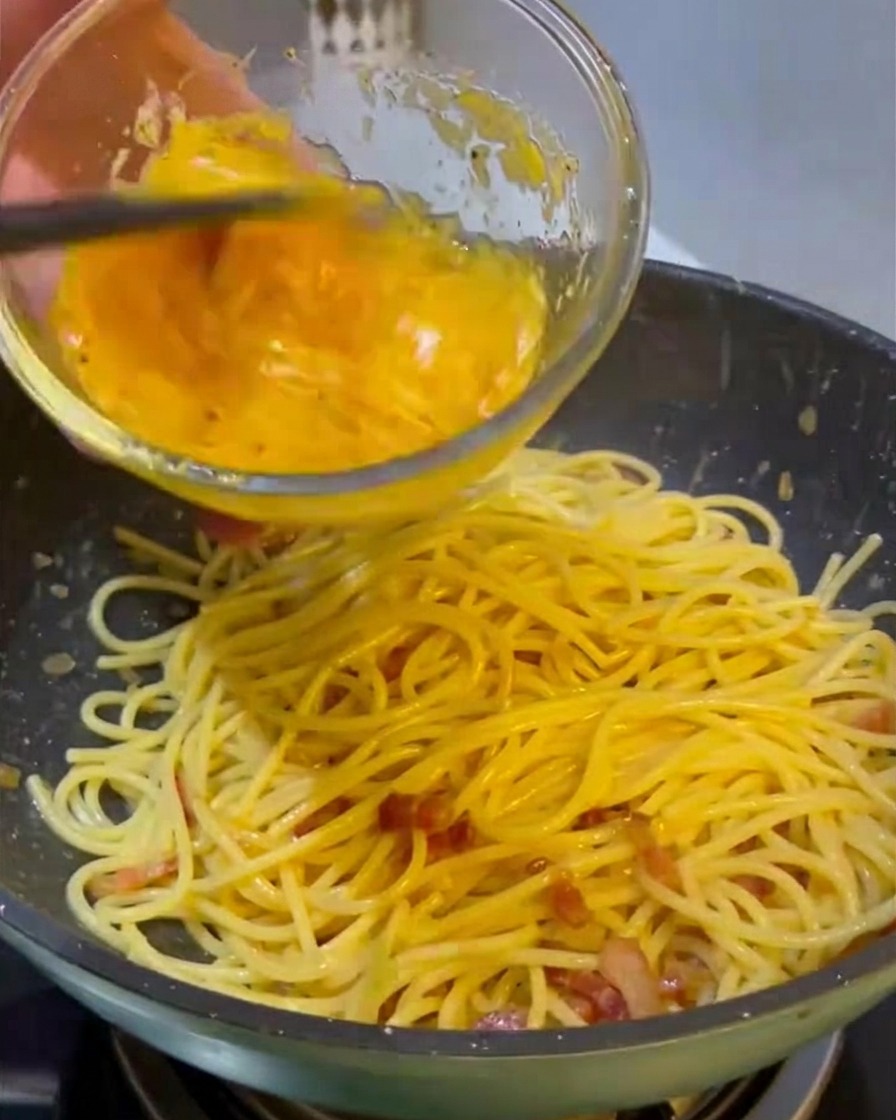 ガラスのボウルから、鮮やかな黄色の卵とチーズのソースを、熱々のスパゲッティと炒めた肉が入ったフライパンに注ぎ込んでいる様子。