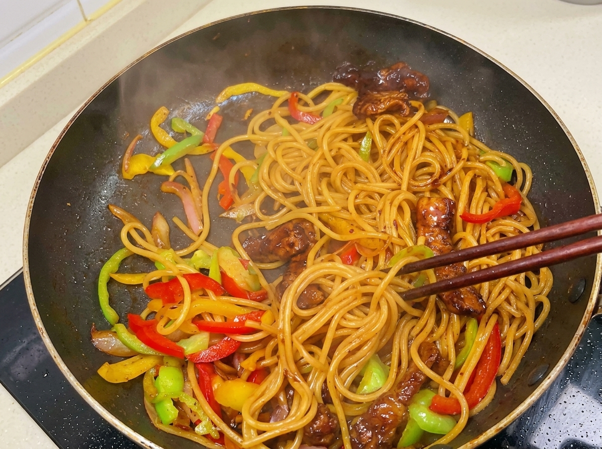 濃いツヤのあるソースが絡んだスパゲティ、香ばしく焼いた豚肉、スライスしたパプリカの湯気立つ混合物を、箸で勢いよく混ぜ合わせている様子。