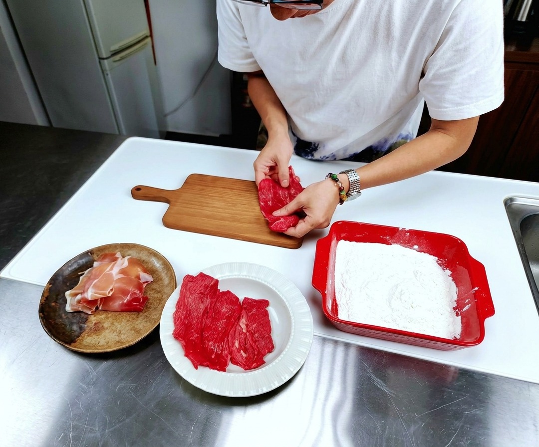 ステンレスの調理台の上で、薄くスライスされた生の仔牛肉が木製のまな板に平らに並べられている様子