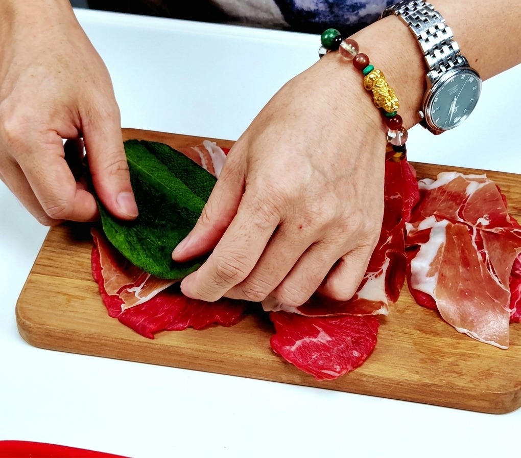 生ハムを重ねた仔牛肉の中央に、手で新鮮な緑色のセージの葉をのせている様子
