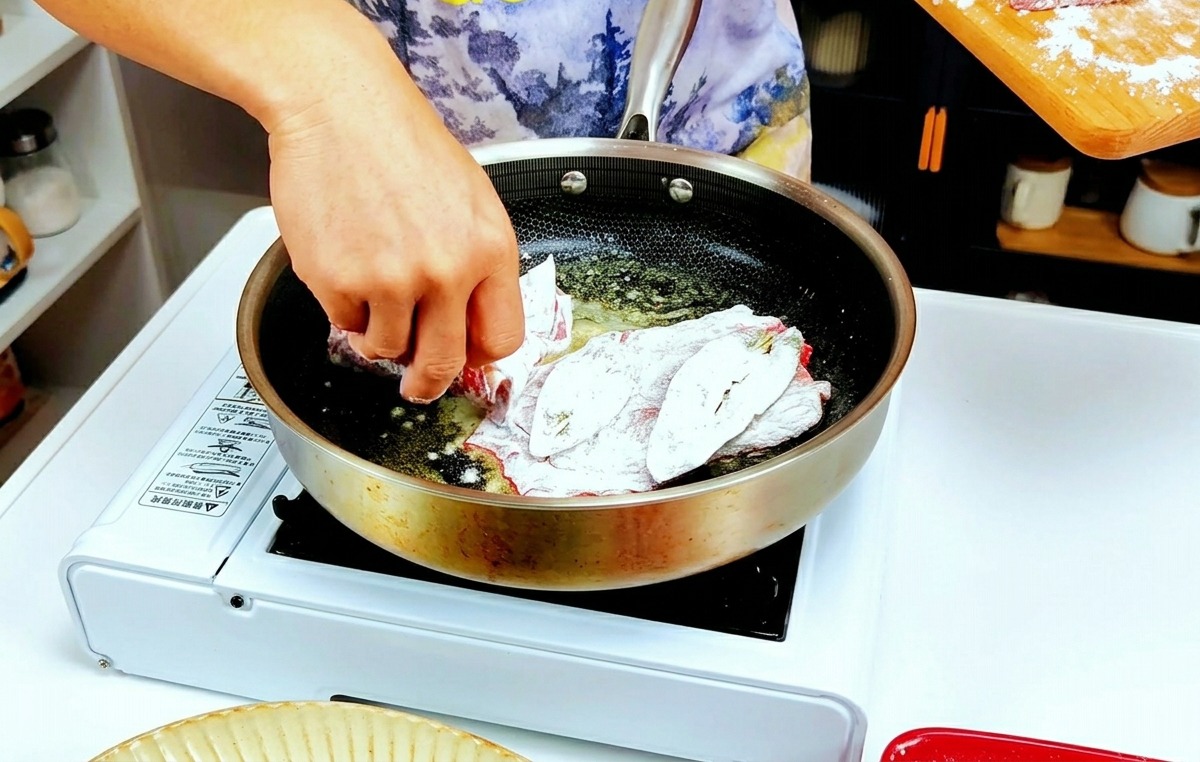 薄力粉を軽くまぶした仔牛肉を、バターが溶けて泡立つ熱いフライパンに並べている様子