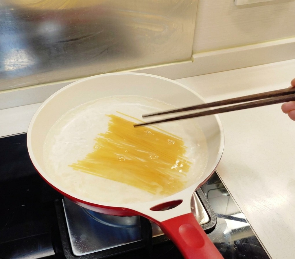 白いフライパンの沸騰したお湯に、木箸で乾燥スパゲティを優しく押し入れている様子。