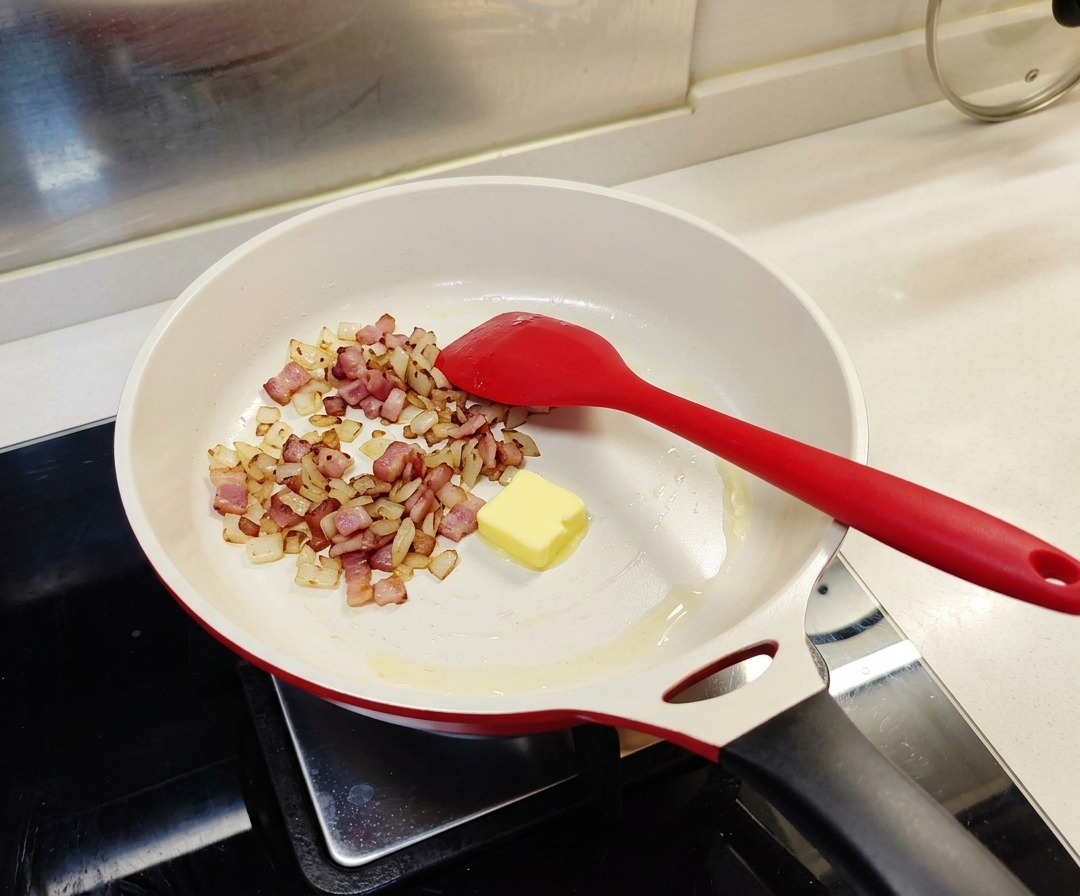 炒めたベーコンと玉ねぎが入った白いフライパンに、四角い黄色いバターが1個入っている様子。