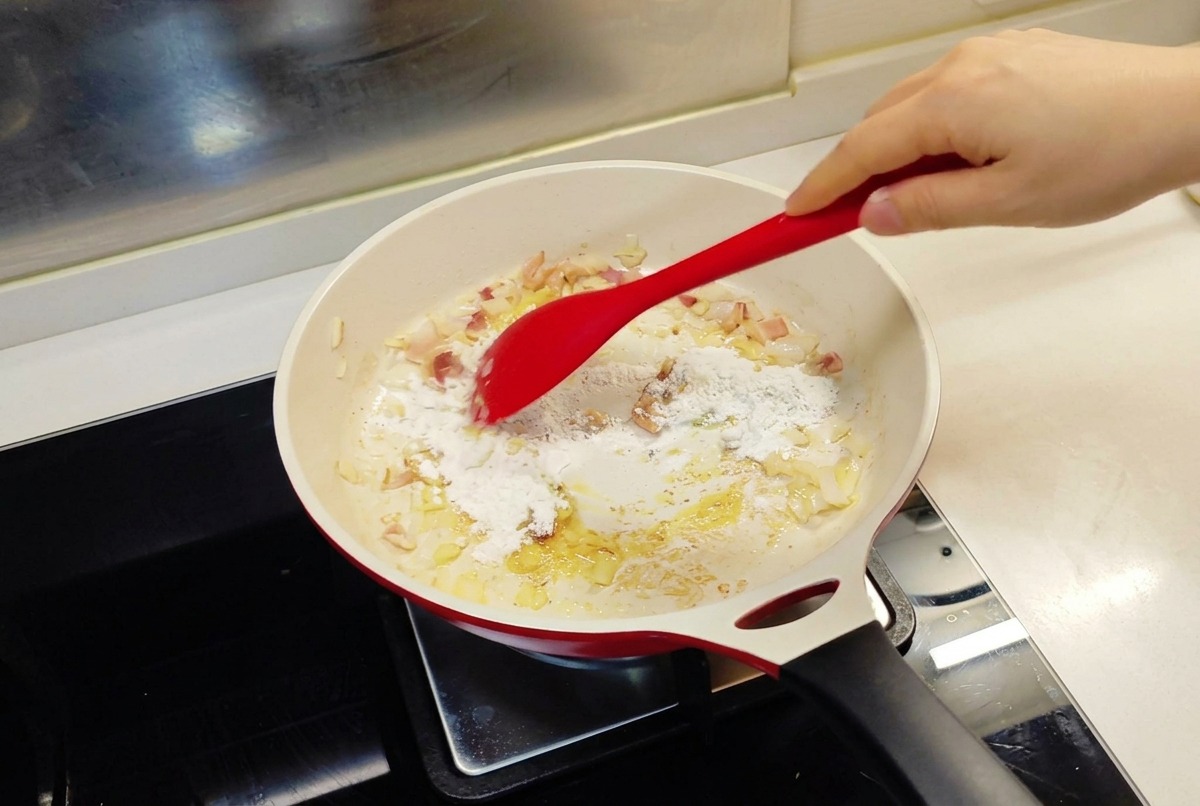炒めた玉ねぎと溶けたバターが入ったフライパンに、赤いスパチュラで薄力粉を混ぜ合わせている様子。