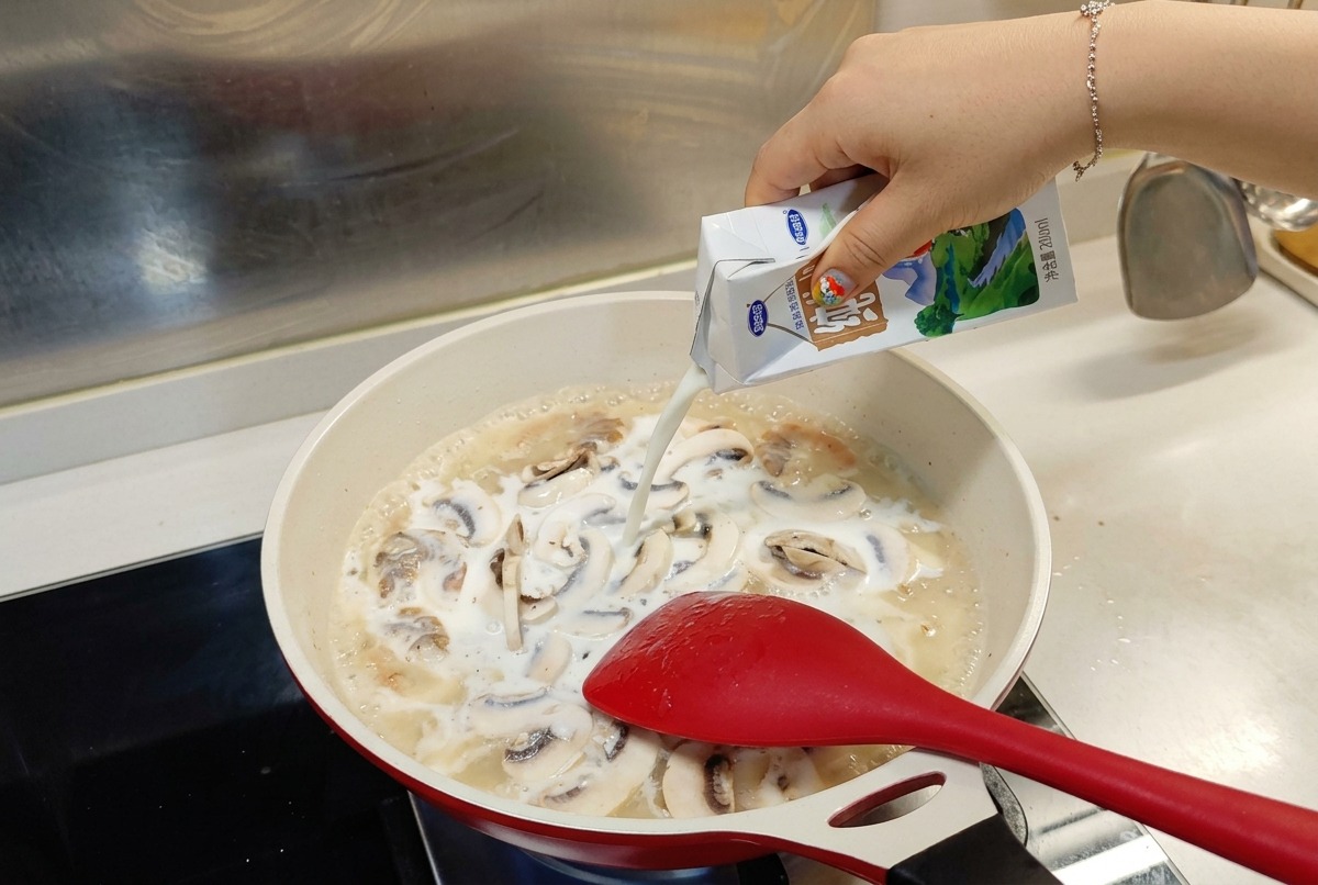 エビとマッシュルームが煮えるフライパンに、白と青のパックから牛乳を注いでいる様子。