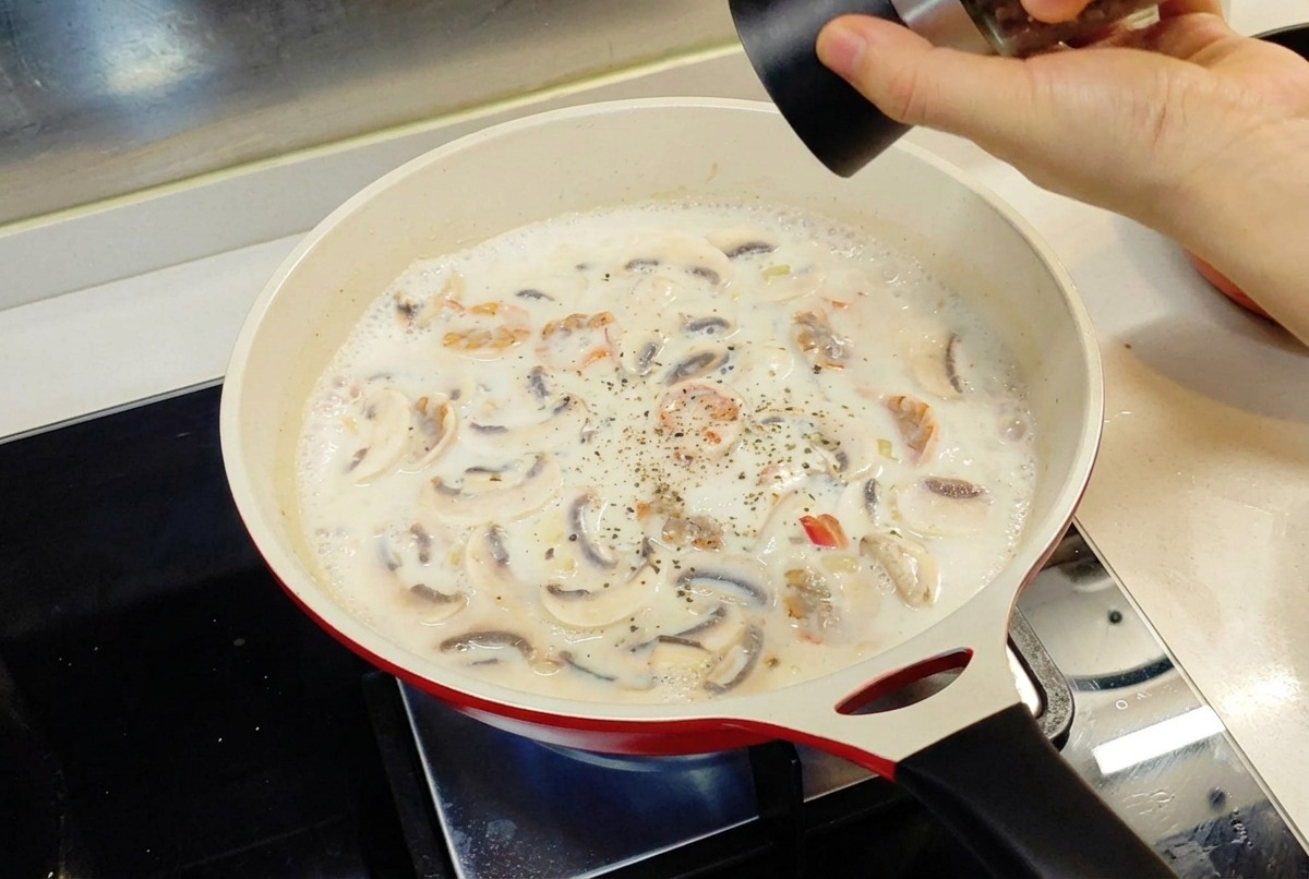 赤いフライパンでグツグツと煮える白いクリームソースに、ペッパーグラインダーで黒コショウを加えている手。