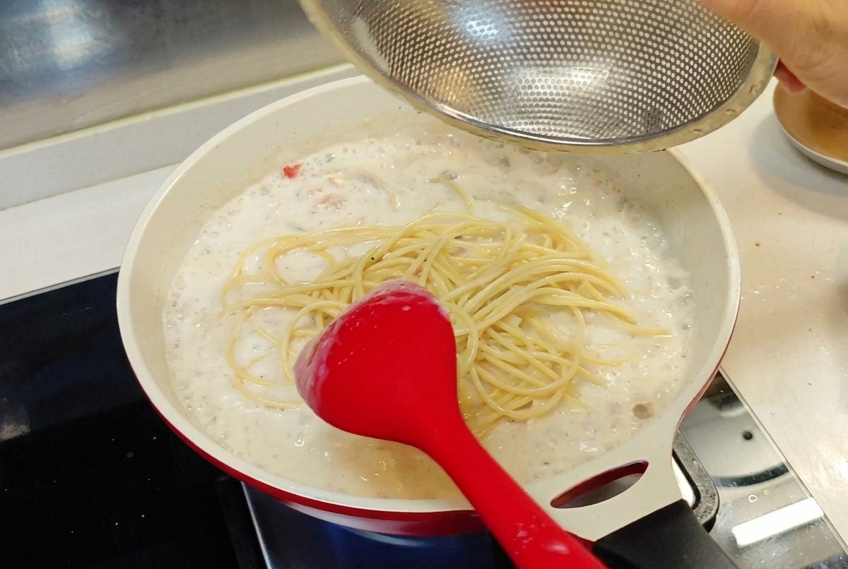 網のザルから、クリーミーなホワイトソースの入ったフライパンに茹で上がったスパゲティを入れている様子。