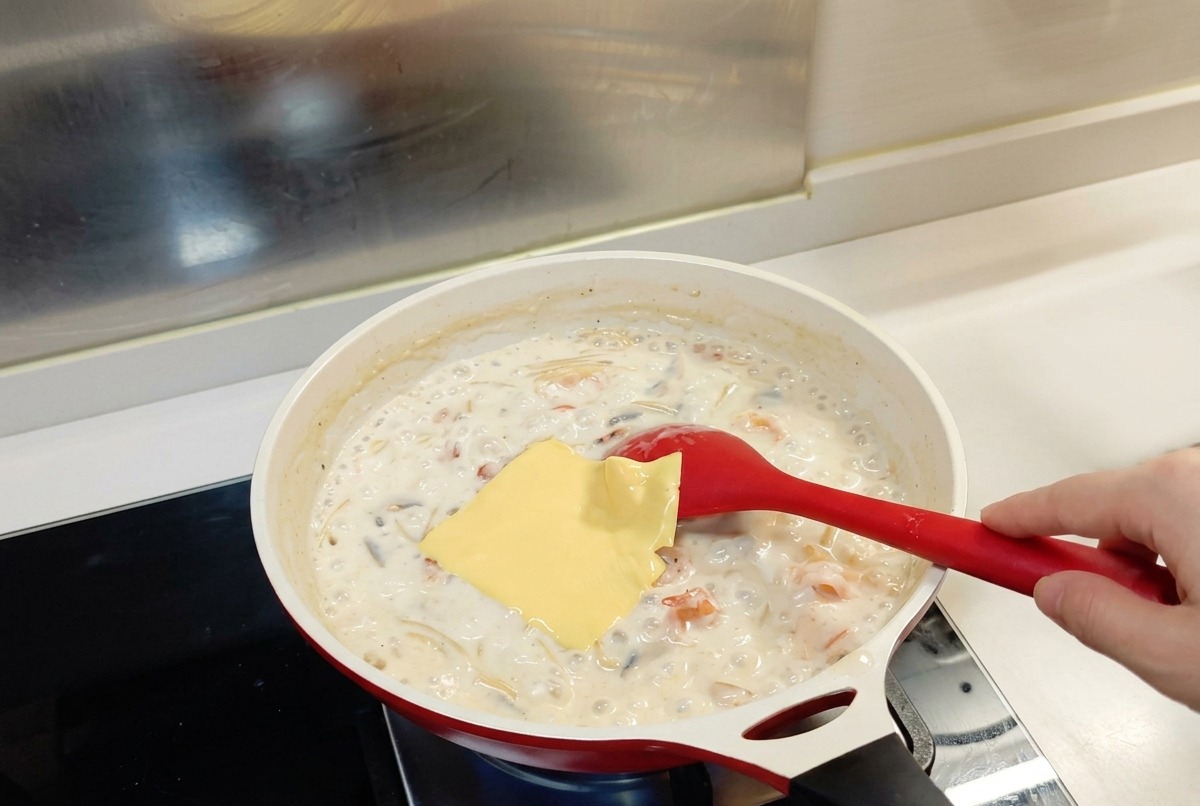 白いフライパンの中でグツグツと煮えるホワイトソースパスタの上に、四角い黄色いチーズが1枚のっている様子。