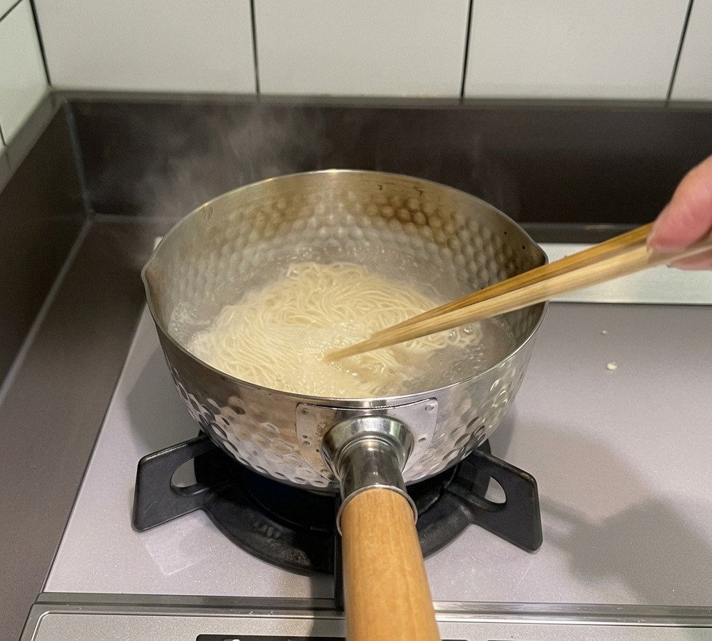 コンロの上の沸騰した鍋で、木箸を使って細い麺を混ぜてほぐしている様子