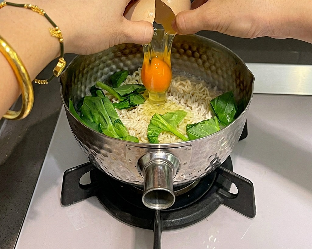茹でているインスタント麺と緑のほうれん草が入った鍋の上で、両手を使って生卵を割り入れている様子