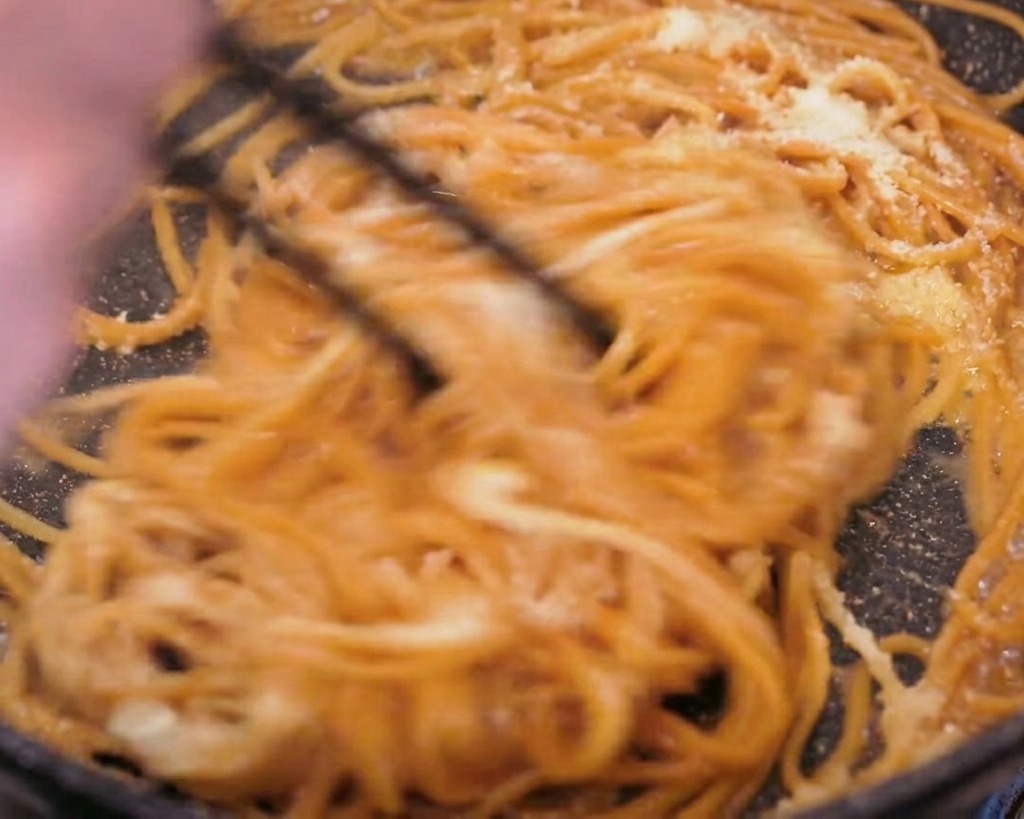 フライパンの中で黒い箸を使い、溶け出すバターと粉チーズをパスタ全体に勢いよく絡めているダイナミックなカット。
