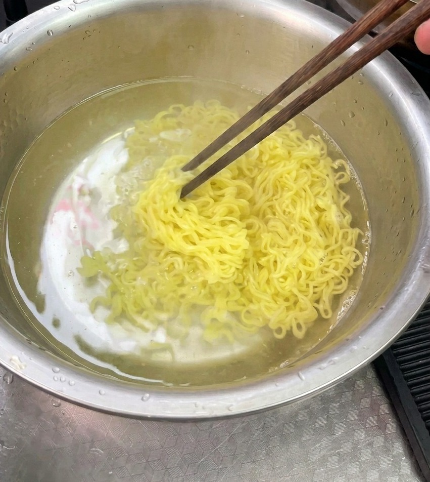 茹で上がった黄色い麺を、ステンレスのボウルに入れた冷水の中で箸を使って洗い流している様子。