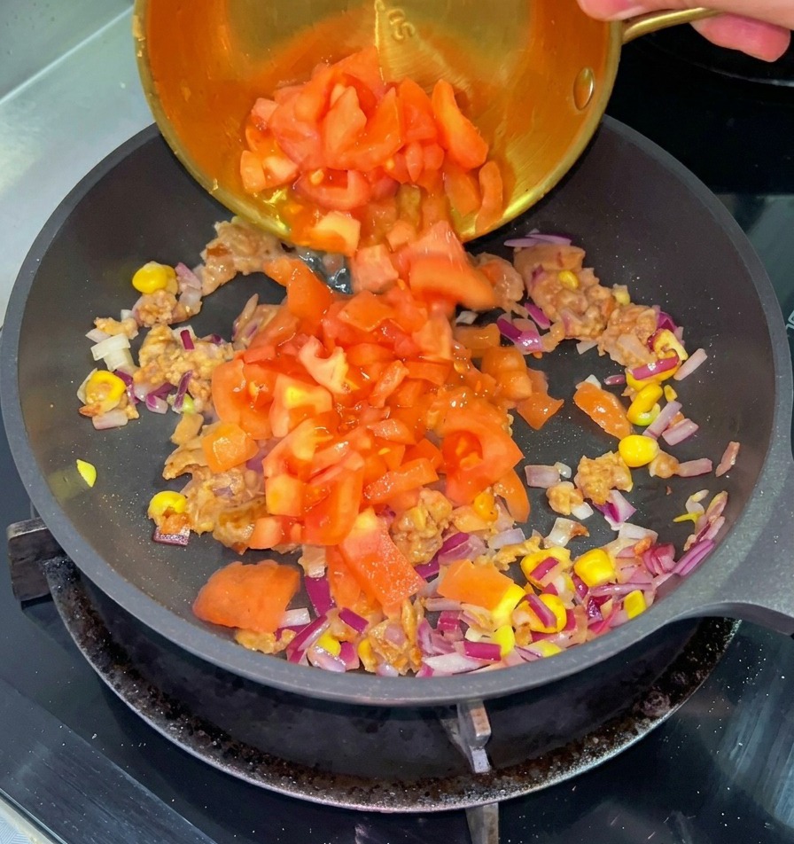 炒めた牛肉、コーン、赤玉ねぎの入ったフライパンに、小さな真鍮のボウルから角切りにした赤トマトを注いでいる様子。