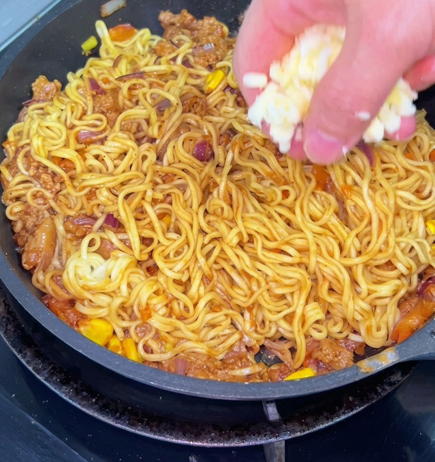 ひき肉、玉ねぎ、コーン、麺が混ざった炒め麺の上に、手でたっぷりの白いシュレッドチーズを散らしている様子。