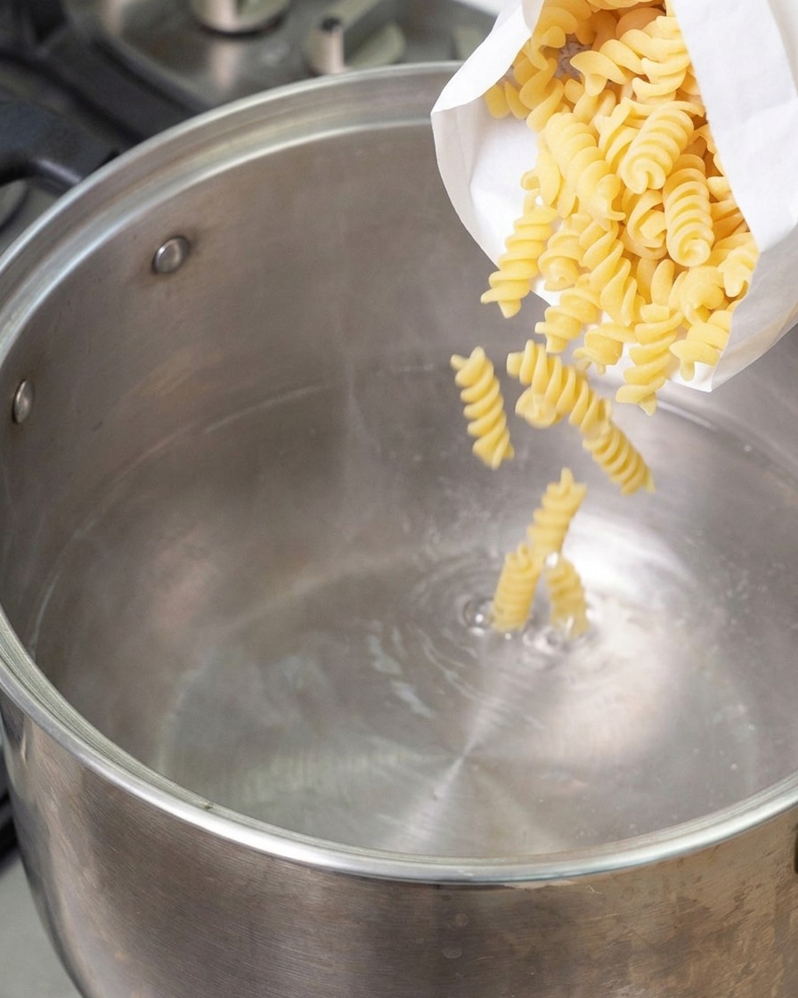 沸騰したステンレスの鍋に、白い袋から乾燥したスパイラル状のフジッリパスタを投入している様子。