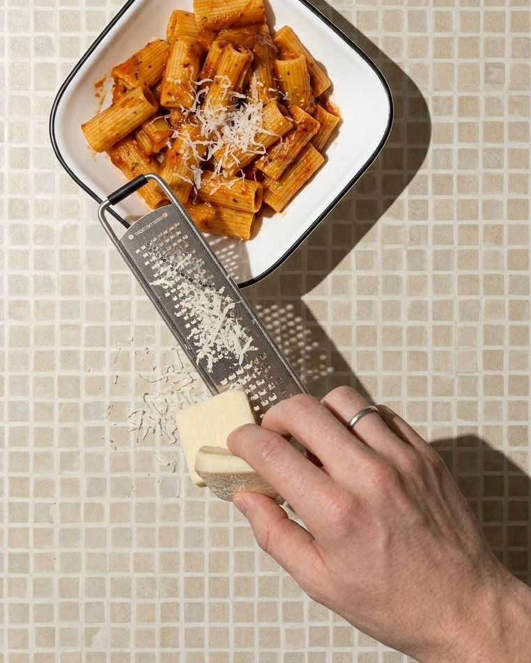 赤いソースを纏ったリガトーニが盛られた白い四角いボウルの上で、手元でブロックのホワイトチーズを削りおろしているクローズアップ。
