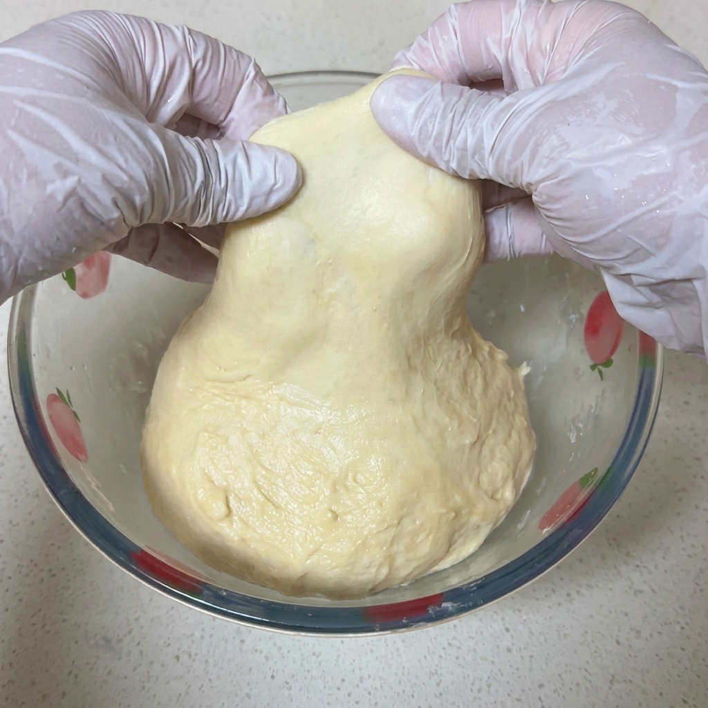 白い手袋をはめた両手でピザ生地をそっと広げ、光に透けるほど薄く破れずに膜が張っている様子を示す様子。