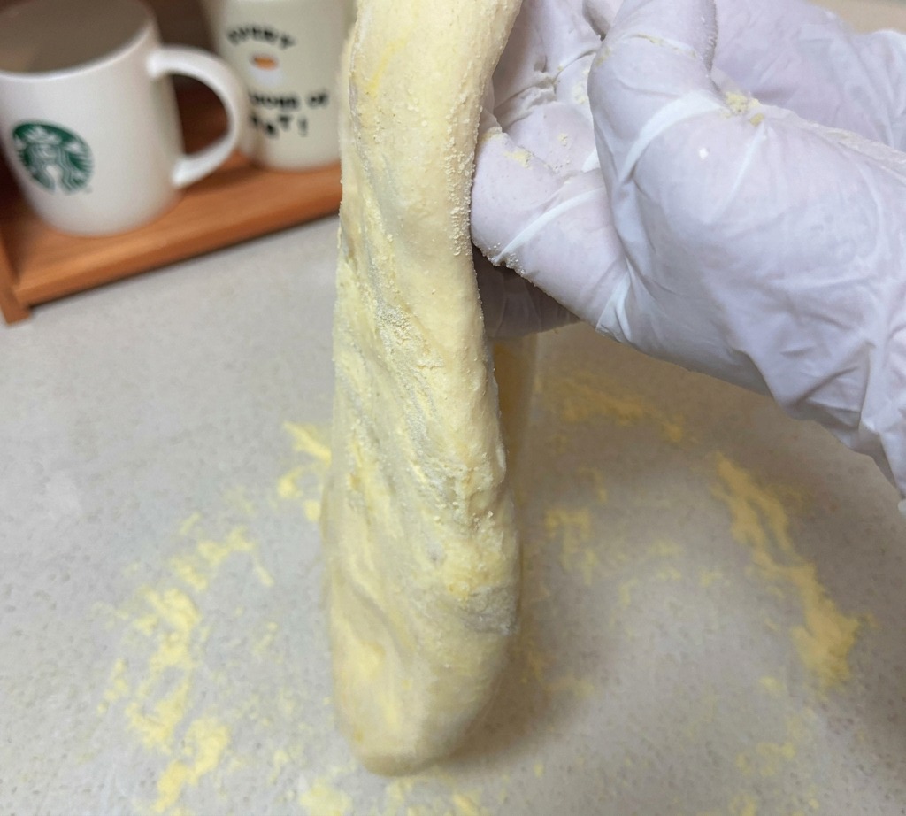 粉が振られた作業台の上で、手袋をはめた両手によって縦に引き伸ばされた生のピザ生地が持ち上げられている様子。