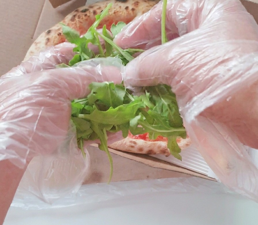 箱の中の焼き上がったピザの中心に、透明なプラスチック手袋をはめた両手が新鮮な緑色のルッコラの葉を山盛りにのせている様子。