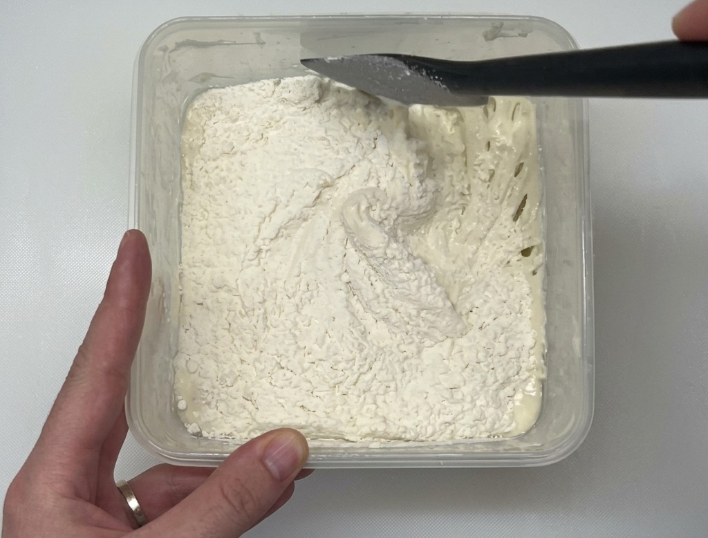 白いワークトップの上の透明なプラスチック容器の中で、黒いスパチュラを使って白い小麦粉を水分多めの生地に混ぜ合わせている様子。
