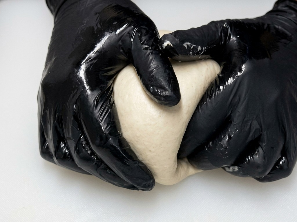 黒い手袋をはめた両手で、ピザ生地を白いまな板の上で転がしながら、なめらかで丸い形に整えている様子。