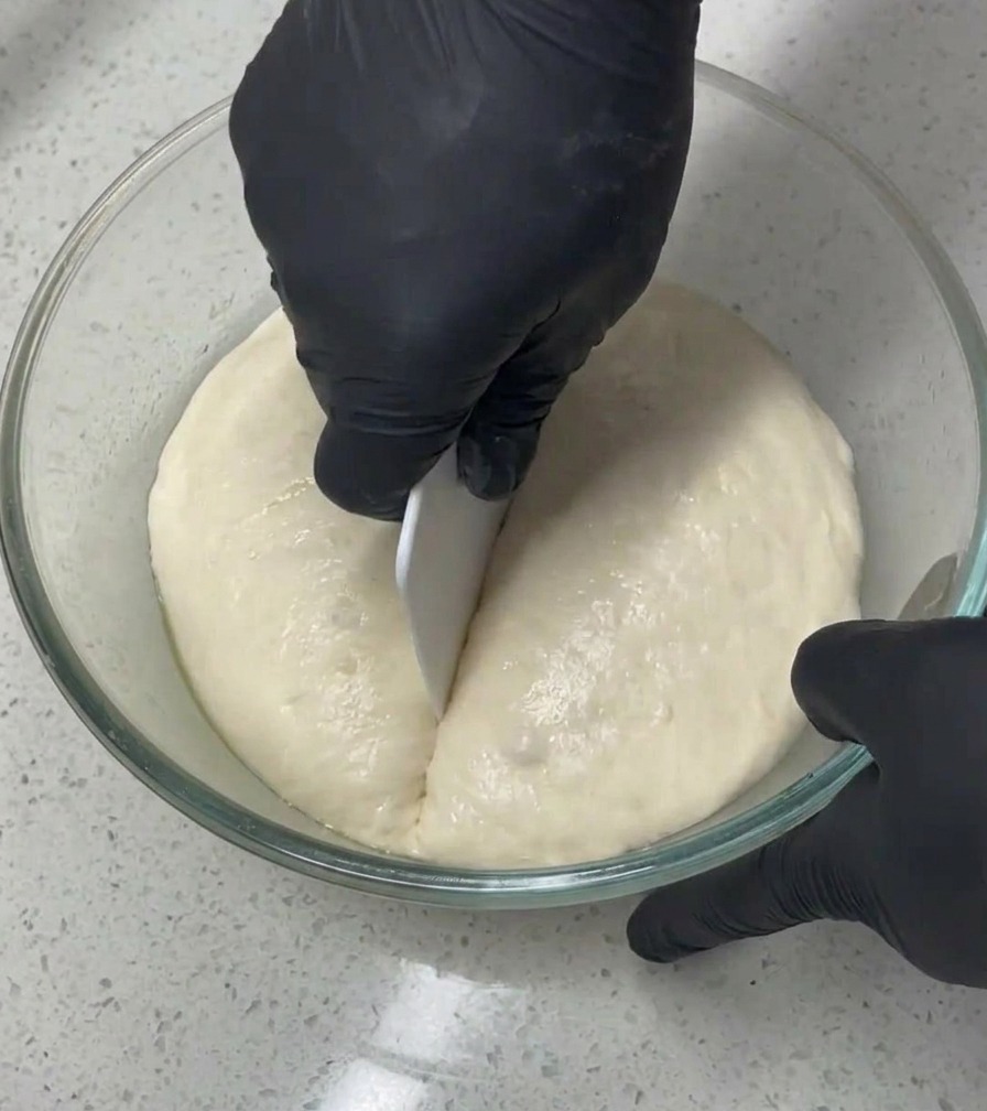 黒い手袋をはめた人が、ガラスボウルの中で白いドレッジを使って、空気を含んで大きく膨らんだ生地を半分に切り分けている様子。