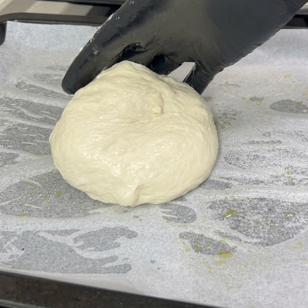 手袋をはめた手が、白いクッキングシートを敷いた天板の上に、柔らかく丸められたピザ生地を置いている様子。
