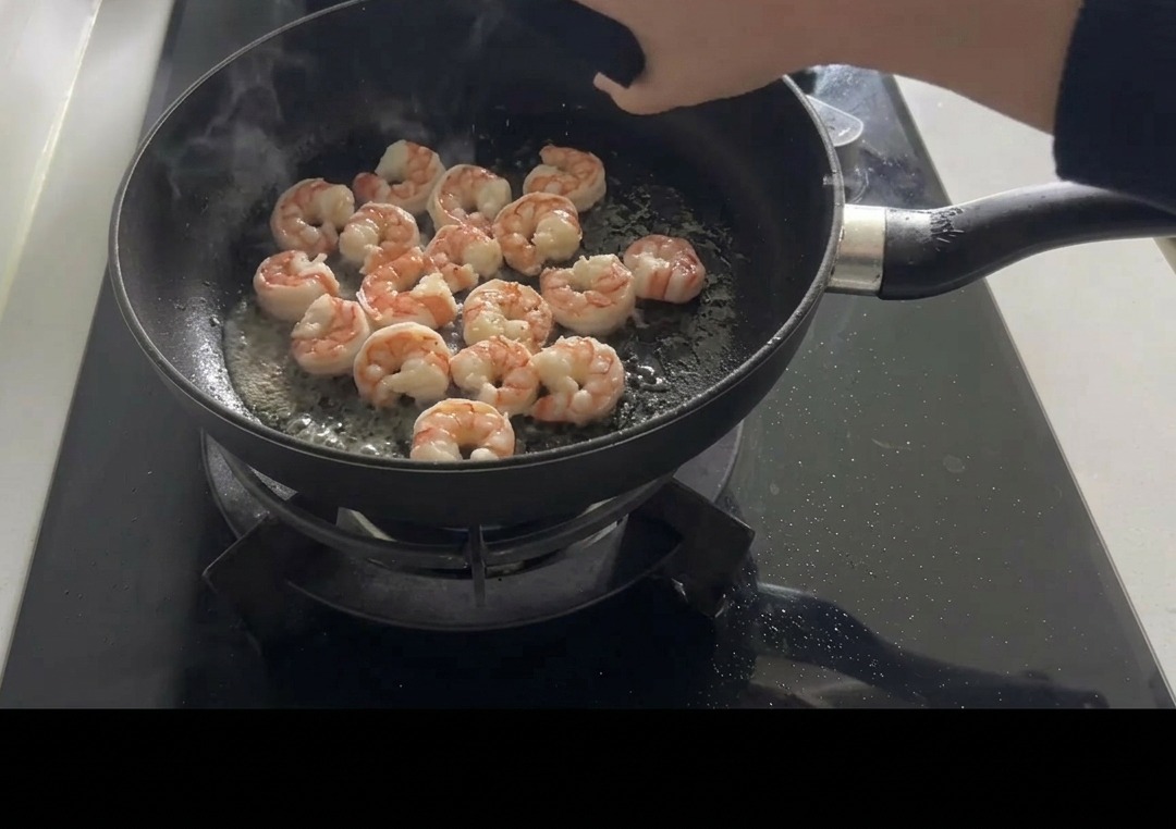 フライパンの中でピンク色に焼き上がったエビに、手で塩とコショウを振って味付けをしている様子。