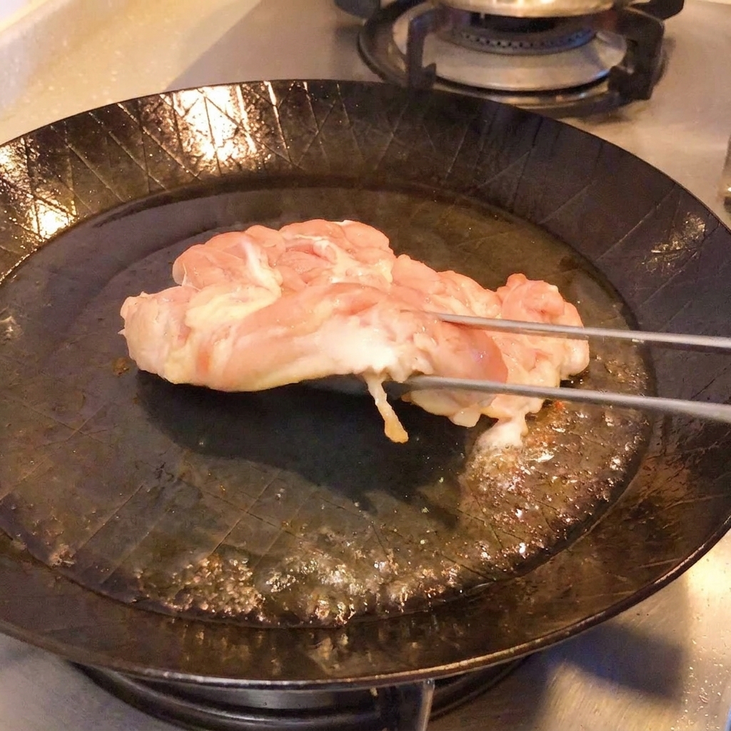 トングでフライパンの鶏肉を持ち上げ、こんがりと黄金色に焼けた皮を見せている様子。