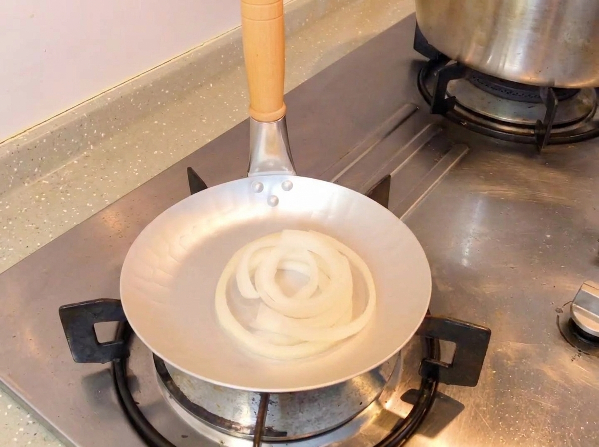 生の玉ねぎが入ったフライパンに、スプーンで濃口醤油のタレを注いでいる様子。