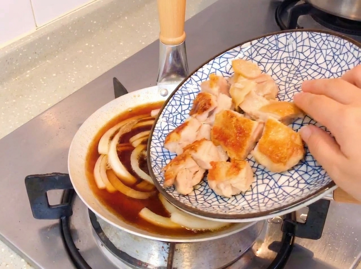 玉ねぎと醤油ベースのタレが煮立つフライパンに、香ばしく焼いた鶏肉の切り身を加えている様子。