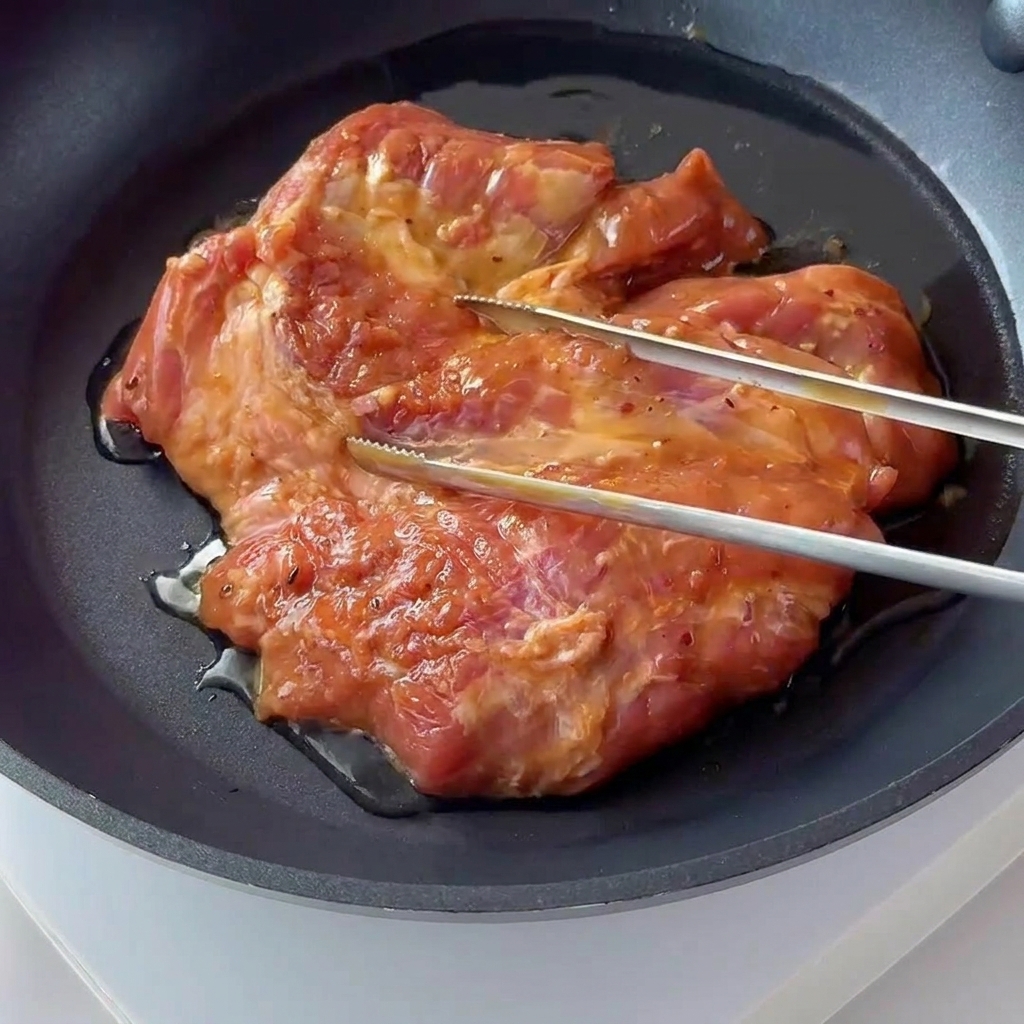 黒い焦げ付き防止フライパンで、味付けしたポークチョップがジュージューと音を立ててこんがりと焼かれている様子。