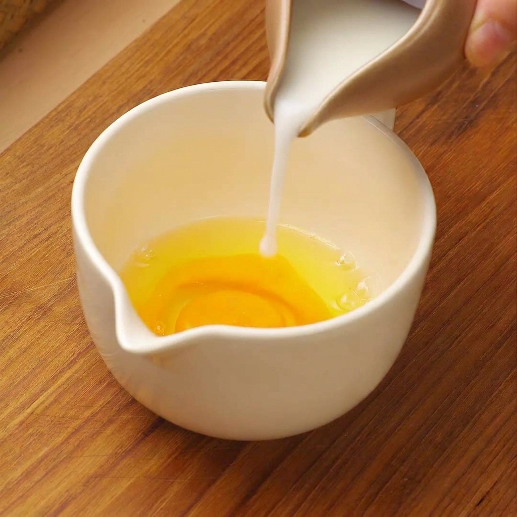 小さなピッチャーから、生卵が入った白い計量カップに牛乳を注いでいる様子。