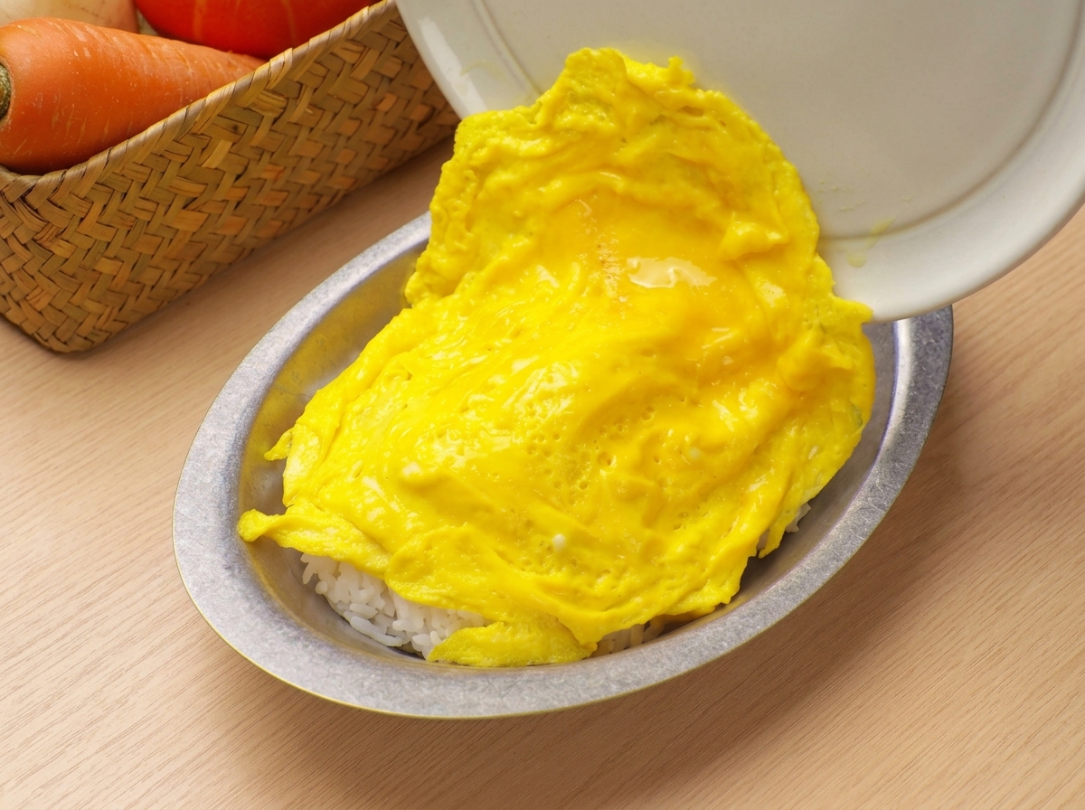 ふんわりとしたスクランブルエッグをフライパンから滑らせて、白いご飯が盛られた器にのせている様子。