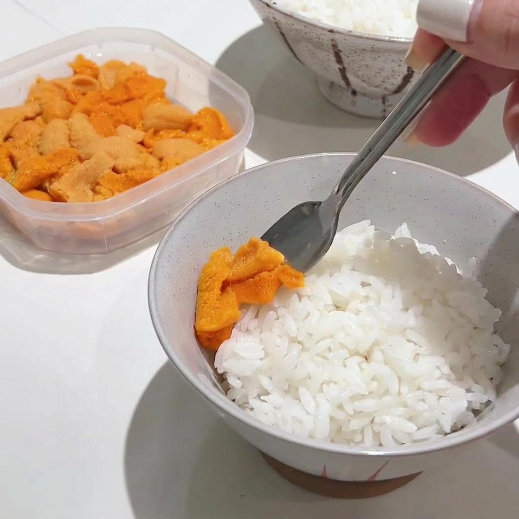 金属製のスプーンを使って、温かいご飯の上に鮮やかなオレンジ色のウニをたっぷりとのせている様子。