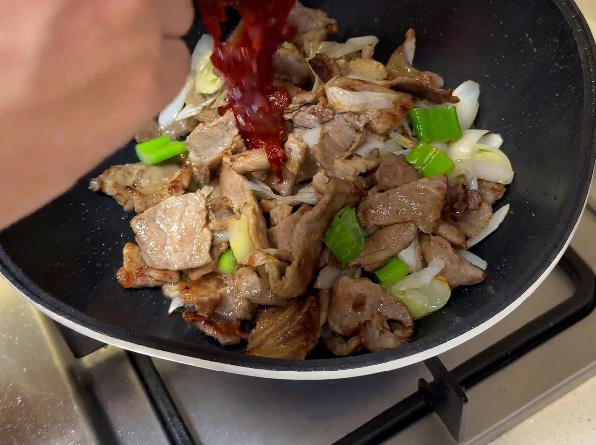 豚肉と長ねぎの上に、小鉢から濃い赤色のタレを流し入れている様子。