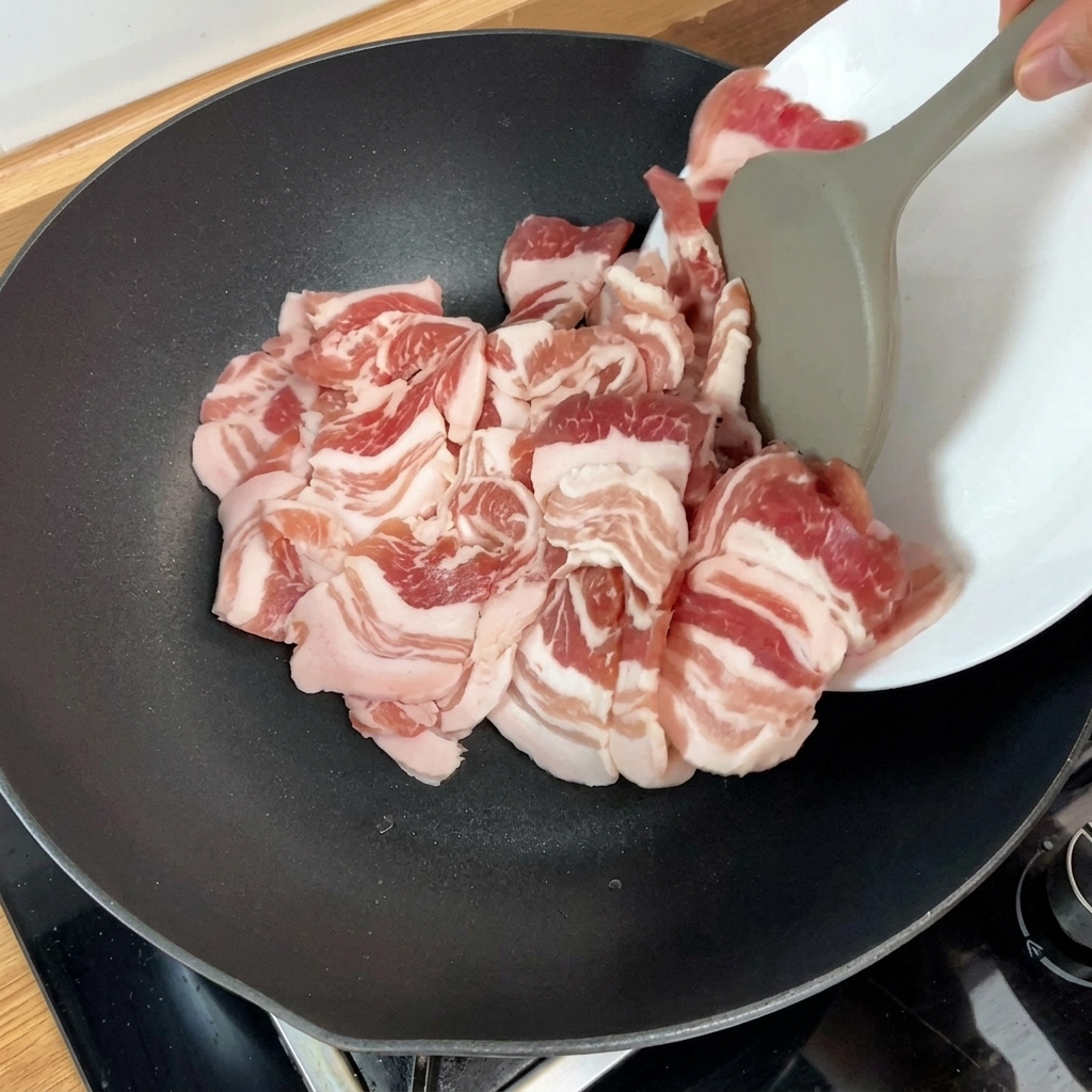 熱したフライパンに調理器具を使って生の豚バラ肉を並べ、焼き始めている様子。