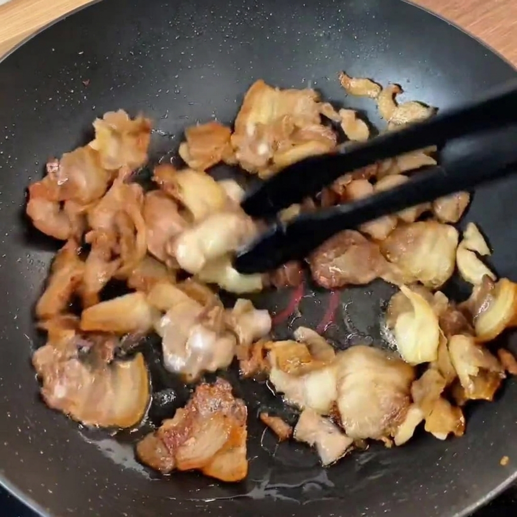 フライパンで焼かれた豚バラ肉の上に、細切りにしたピーマンと紫玉ねぎがのせられている様子。