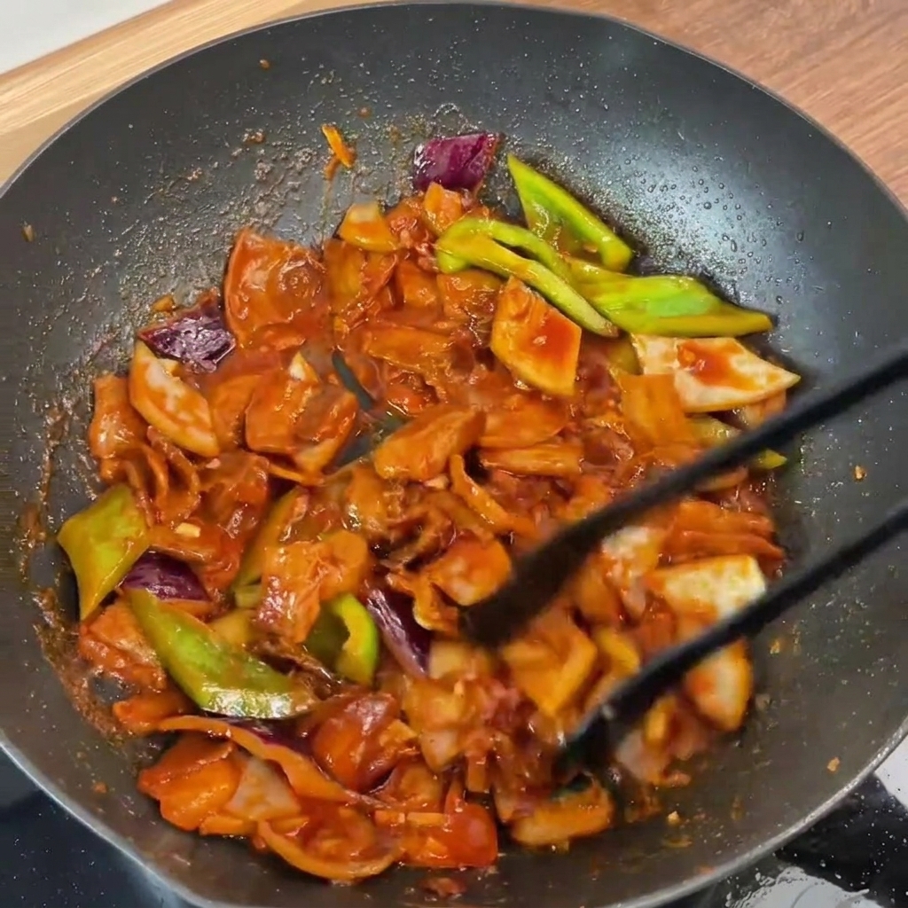 トングを使ってフライパンの中で炒め合わされた、ツヤのある赤いタレが完璧に絡んだ豚バラ肉と野菜。
