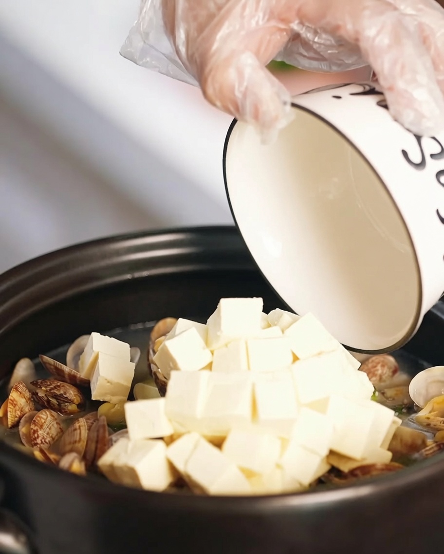 土鍋のあさりスープに角切りの絹ごし豆腐と黄金色の焼き卵を加えている様子